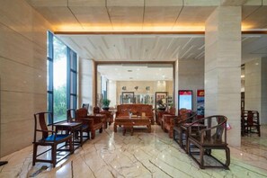 Un vestíbulo amplio con suelo de madera, una zona de estar central con sillas y mesas, y ventanas grandes que dejan entrar luz natural.