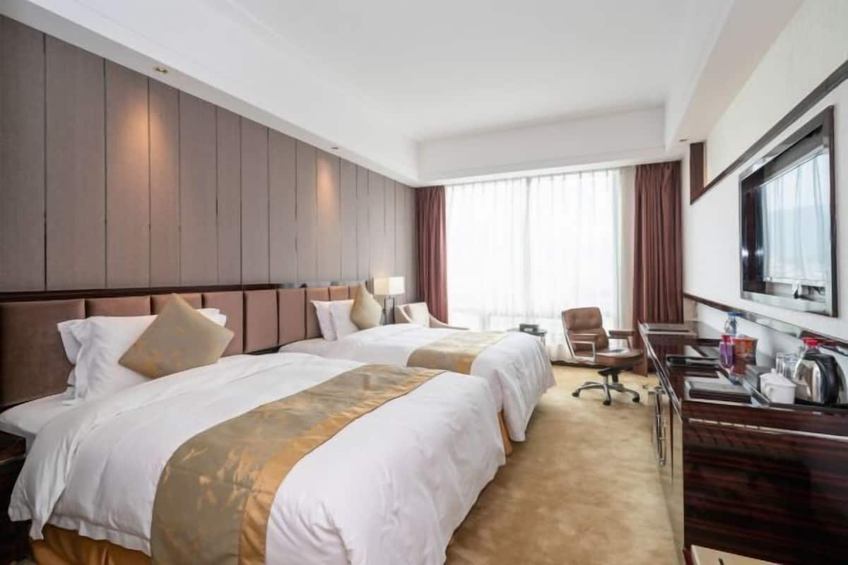 A hotel room with two beds, a desk, a chair, a TV, and a large window with curtains.