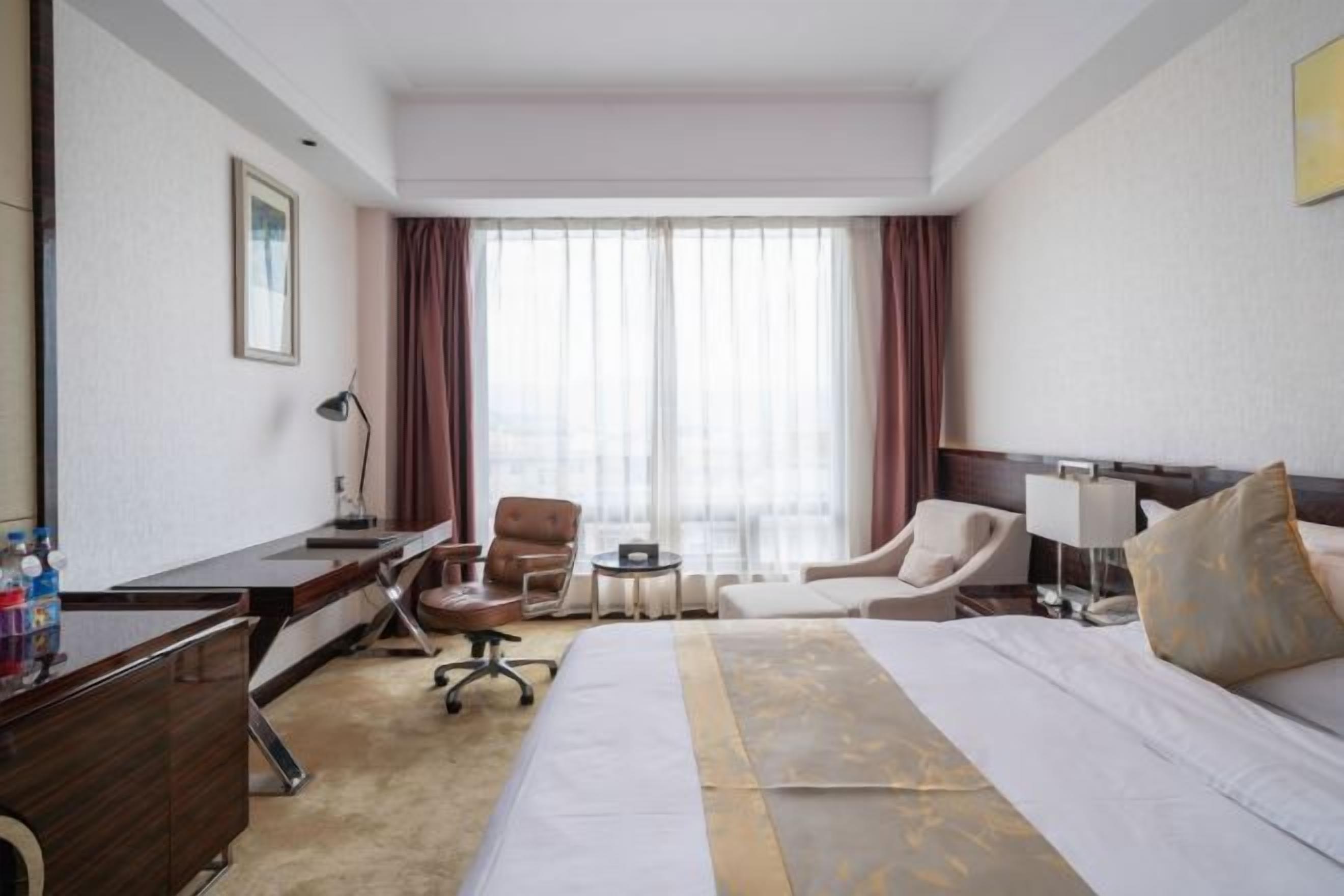 A hotel room with a bed, desk, chairs, a small table, and a window with curtains.