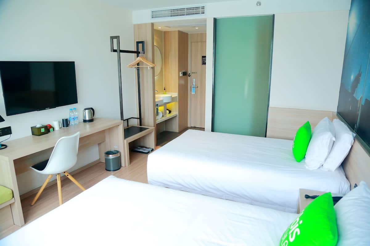 Ein Hotelzimmer mit zwei Betten, einem Schreibtisch mit Stuhl, einem Fernseher und einem durchgegangenen Tür sichtbar liegenden Badezimmer.