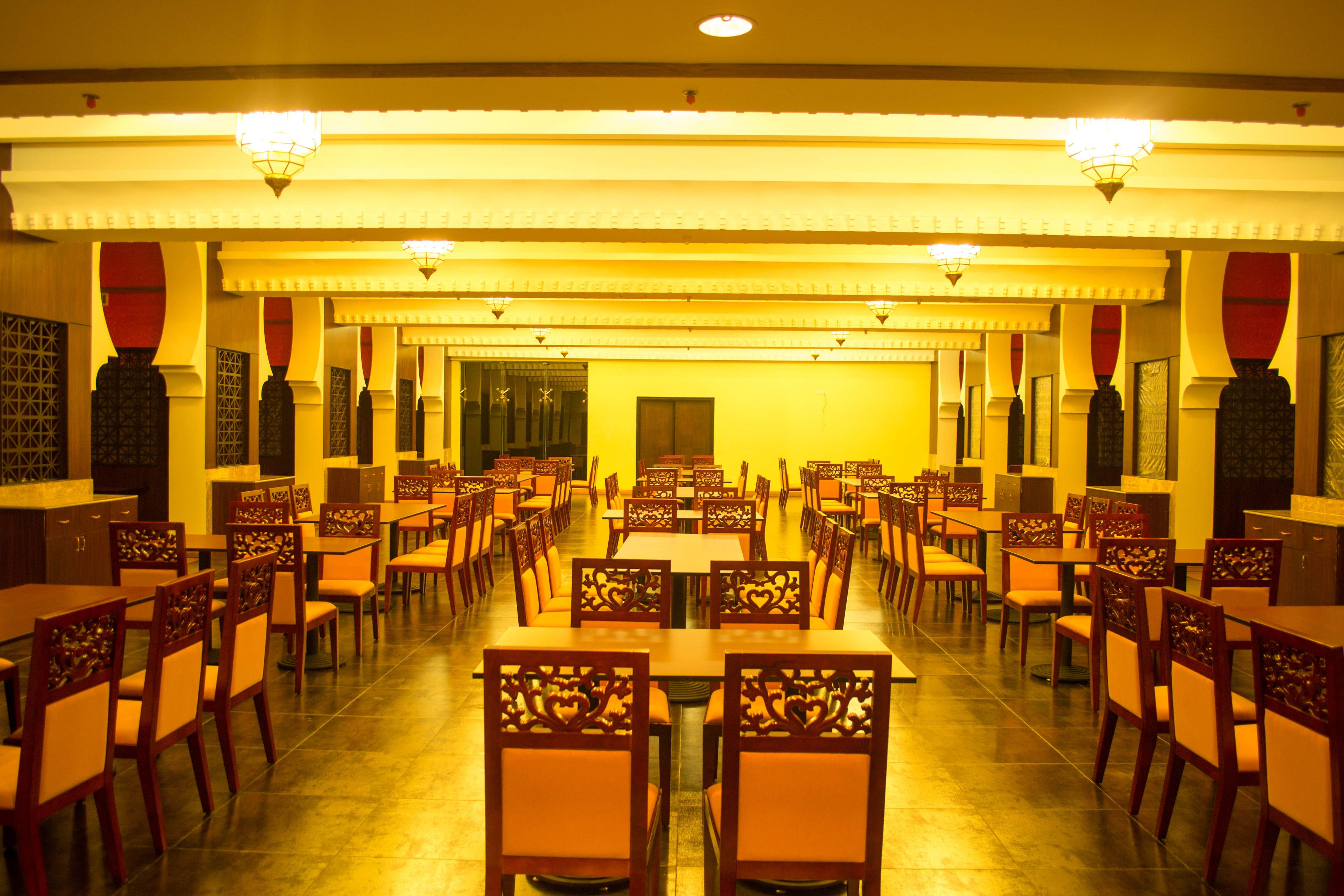 A spacious dining hall with multiple tables and chairs, featuring intricate wooden designs on the chairs and a warm, yellow-lit ambiance.
