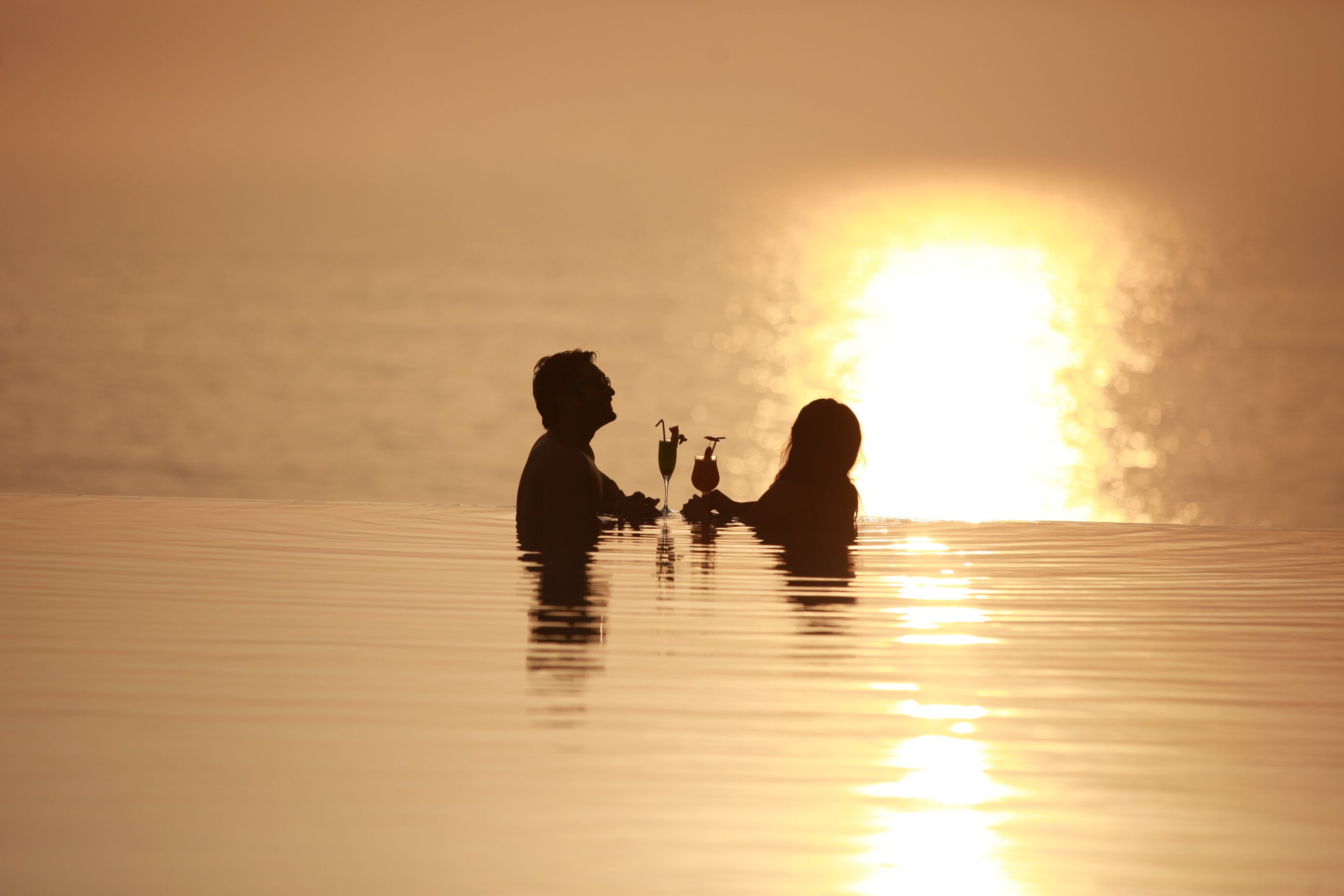On aperçoit la silhouette de deux personnes assises à une table, un verre à la main, face à un coucher de soleil sur l’eau.