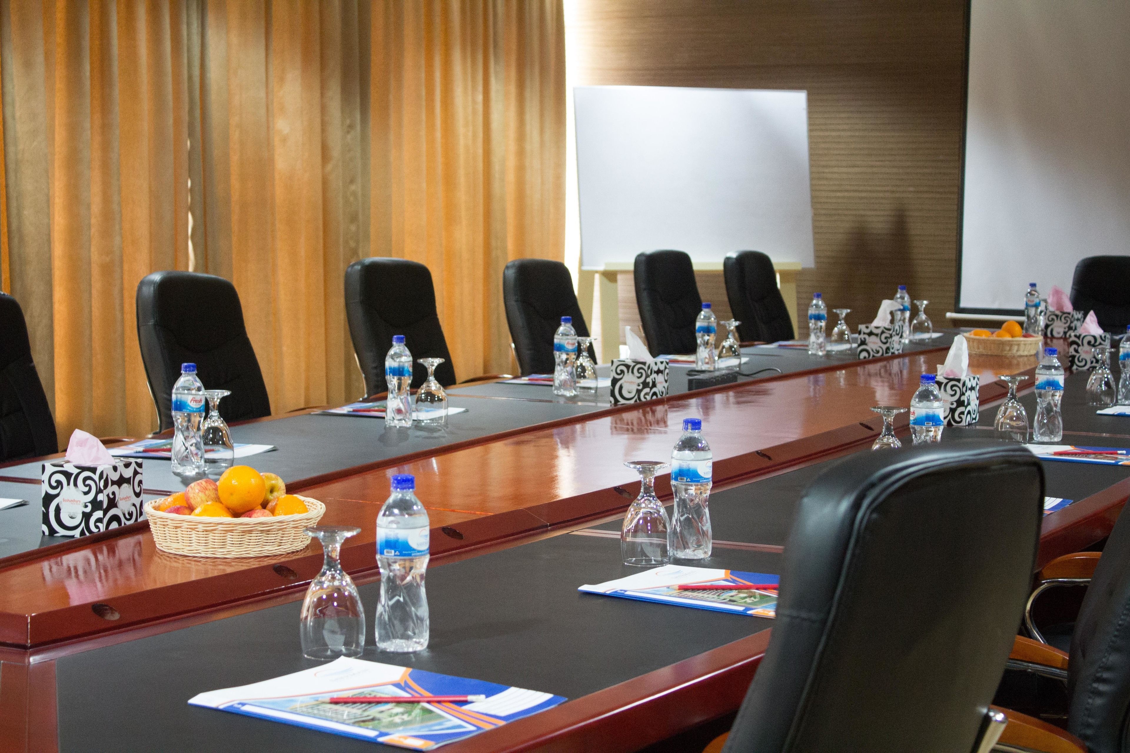 Un salón de conferencias con una larga mesa, sillas, botellas de agua, una cesta de fruta y una pantalla para proyector.