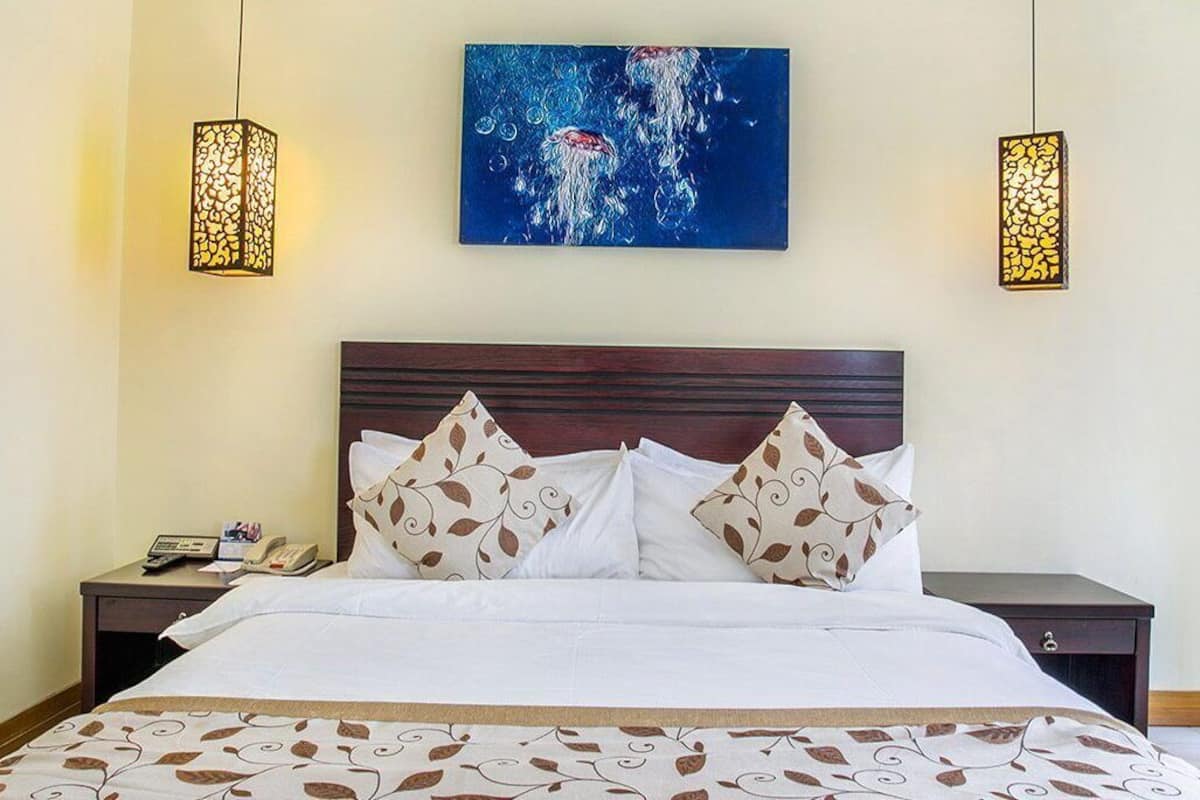 A neatly made bed with patterned pillows and a decorative bedspread, a bedside table with a phone and a lamp, and a painting on the wall.