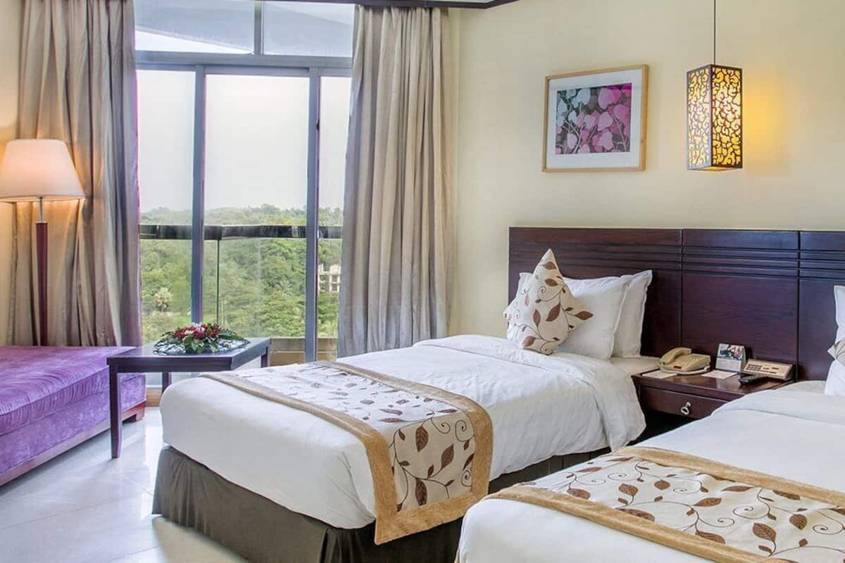 A hotel room with two beds, a desk, a chair, and a window with a view of greenery.