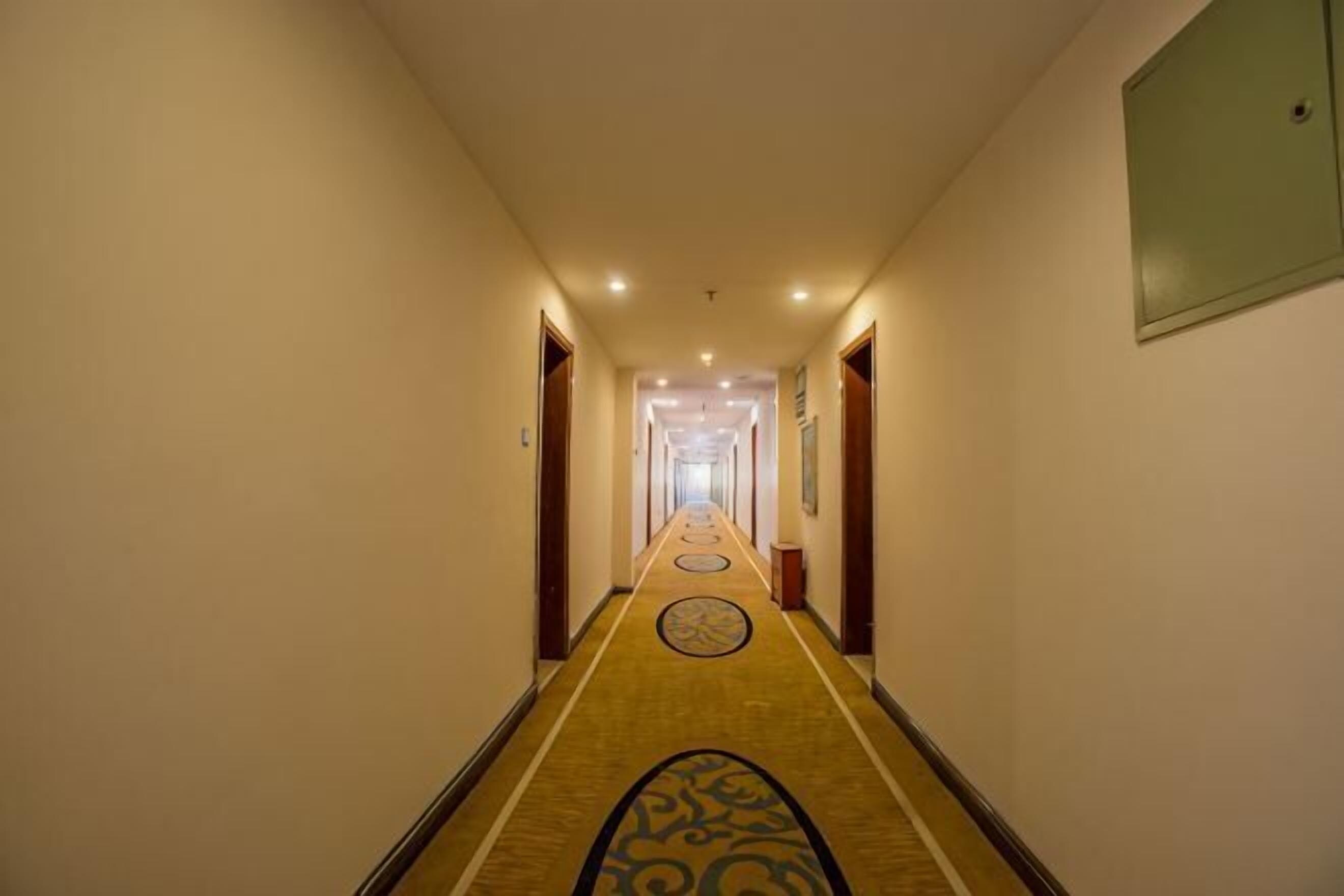 Un couloir d’hôtel avec un sol recouvert de moquette, des portes de chaque côté et un luminaire au plafond.