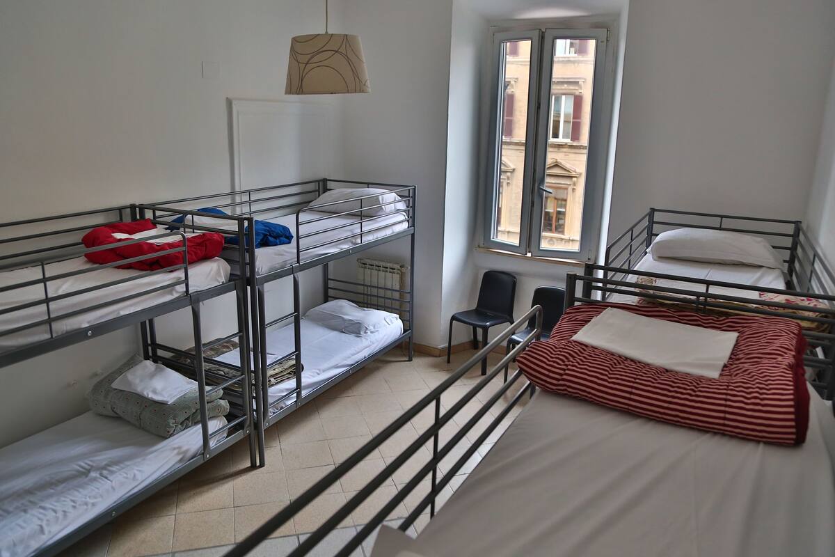 A dormitory room with bunk beds, a window with a view of buildings, and a chair.