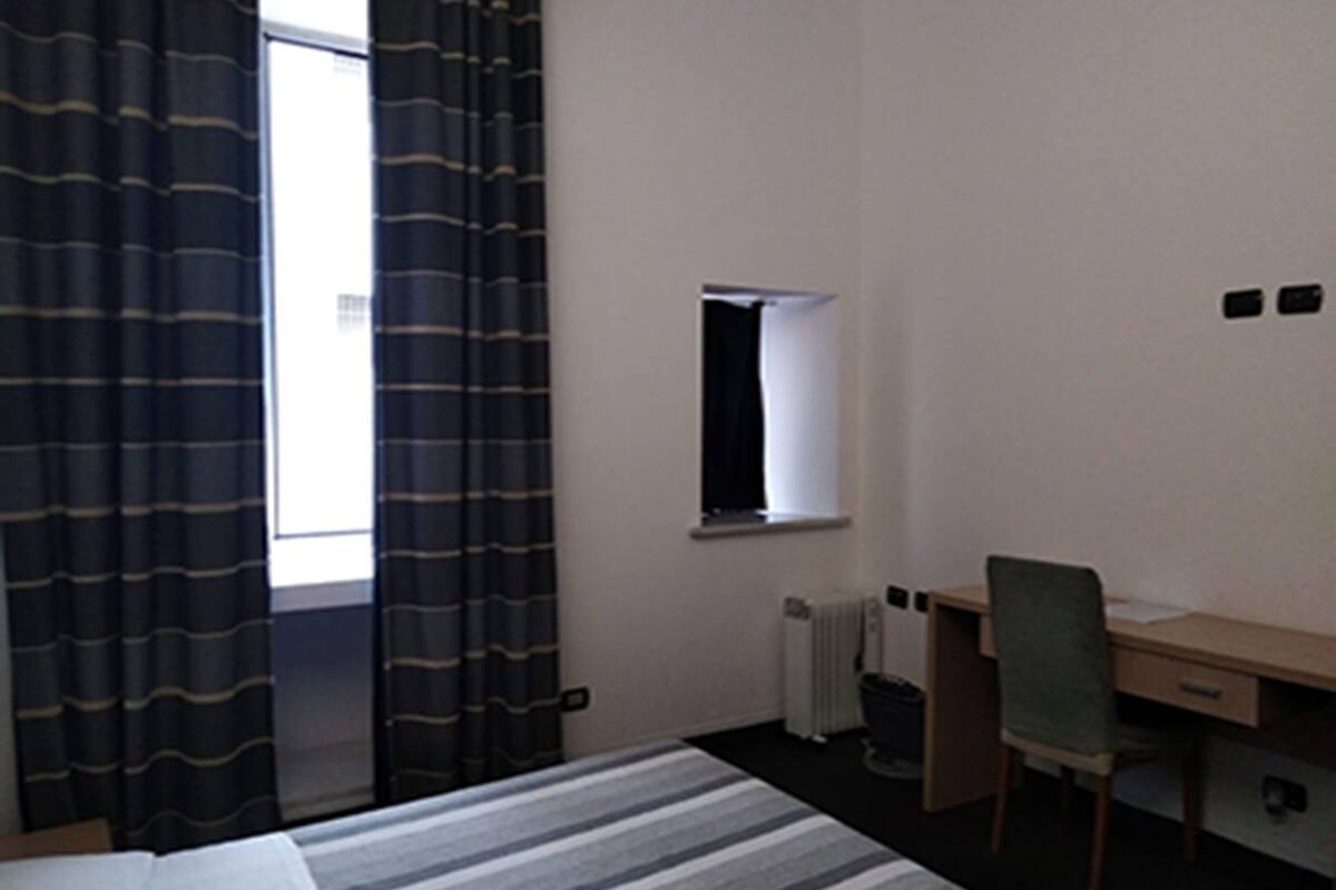 A hotel room with a bed, a desk, a chair, a radiator, and a window with curtains.