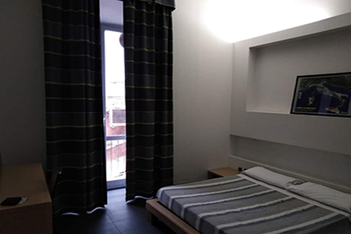 A small, dimly lit room with a single bed, a nightstand, and a wall-mounted shelf.
