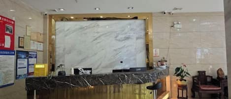A hotel reception area with a marble reception desk, a seating area with chairs, and a wall-mounted information board.