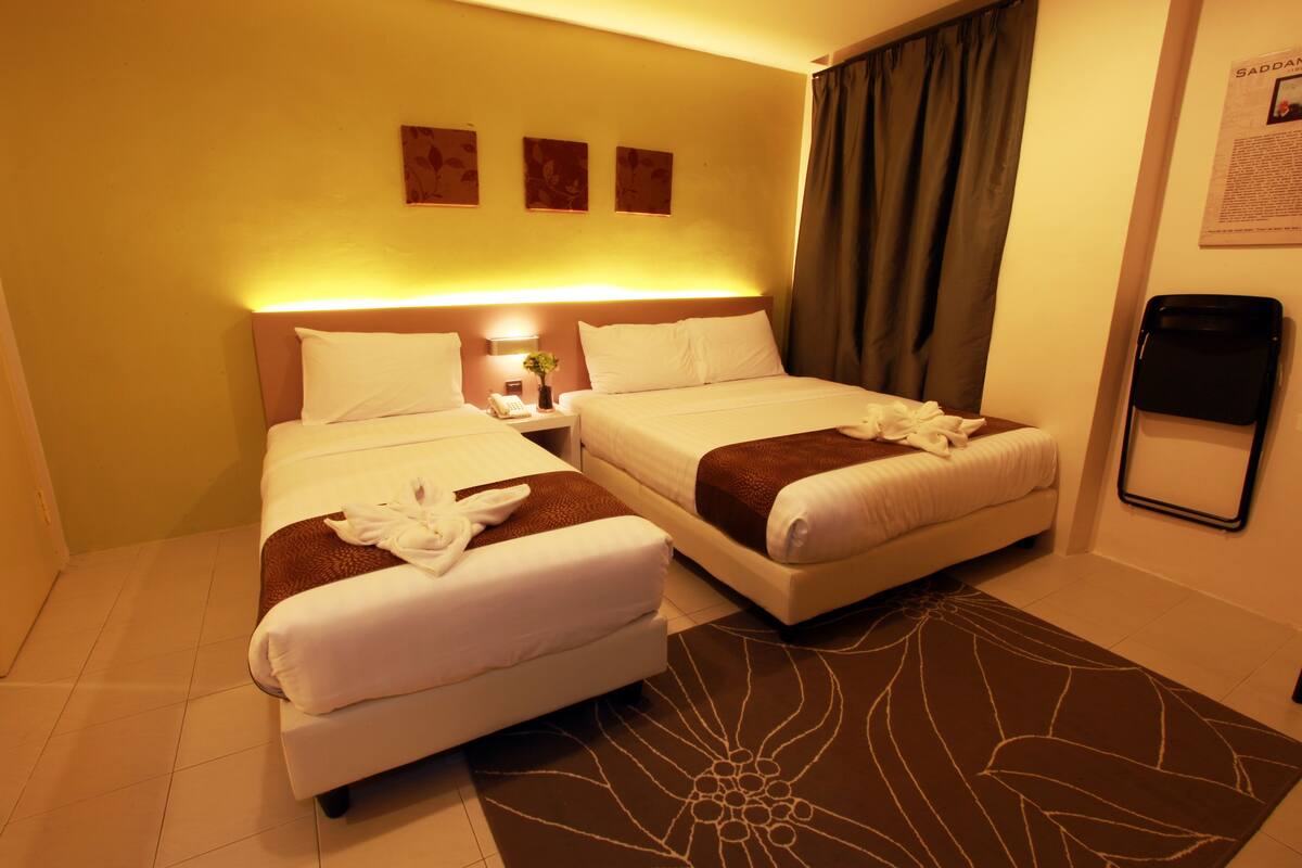 Habitación de hotel con dos camas, una mesita de noche, una lámpara y una alfombra marrón con estampado floral.