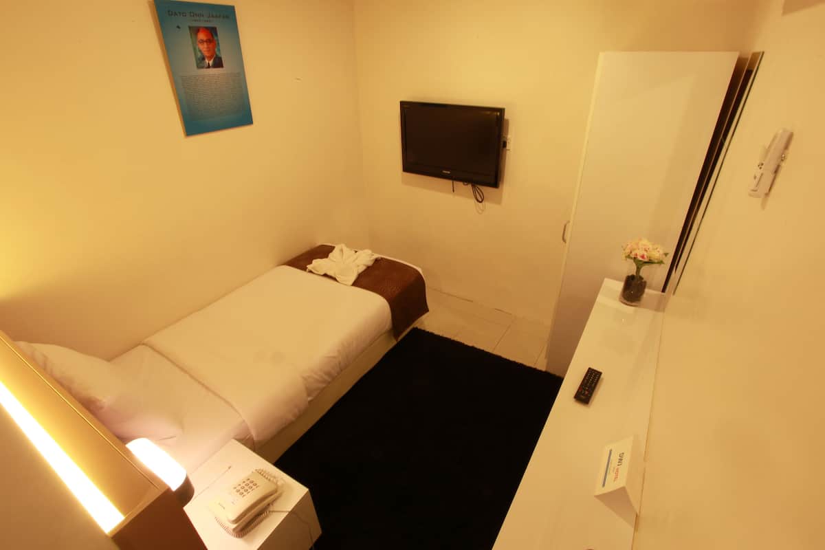 Una habitación de hotel pequeña y bien iluminada con una cama individual, una mesita de noche con lámpara, un escritorio con teléfono, un televisor de pantalla plana montado en la pared y un espejo en la pared opuesta.