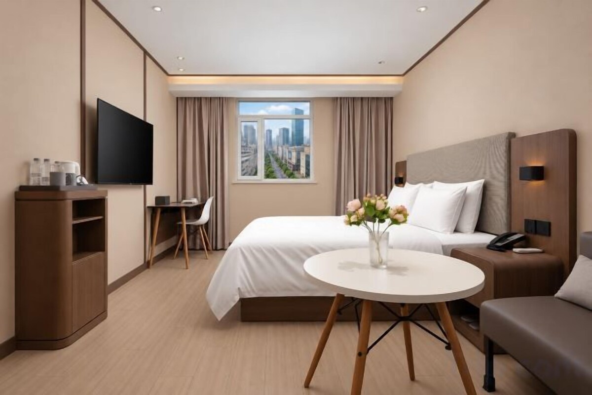 Habitación de hotel con cama, escritorio, televisión, ventana con vista a la ciudad y un jarrón con flores.