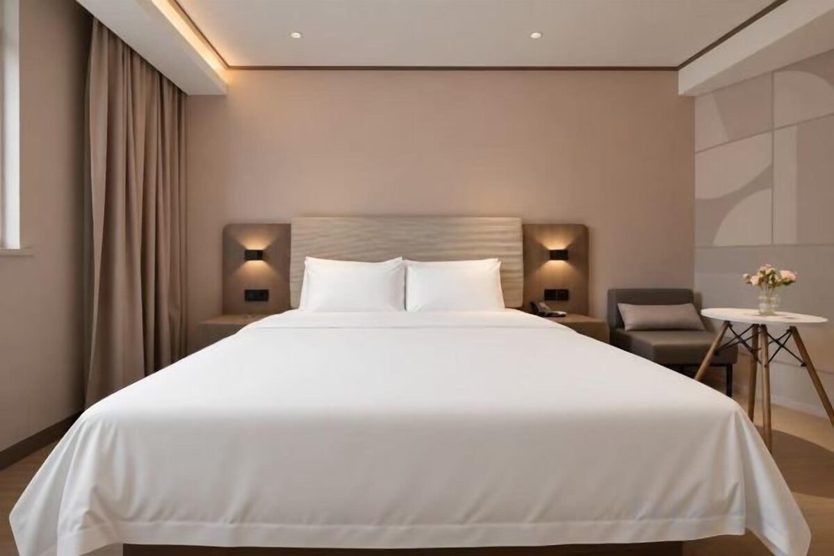 Habitación de hotel con una cama grande, mesitas de noche, una silla, una pequeña mesa con flores y una ventana con cortinas.