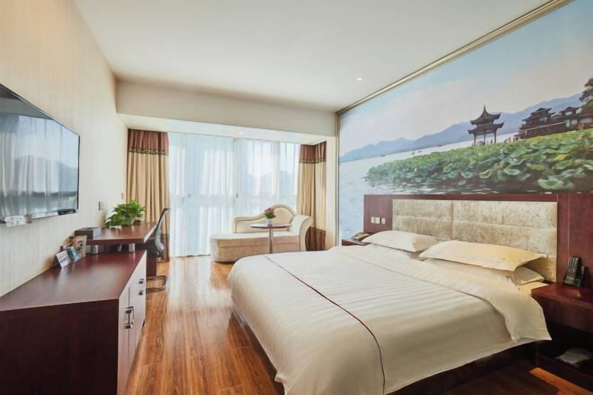 A hotel room with a large bed, a desk with a chair, a small table with a plant, and a view of a traditional building through a window.
