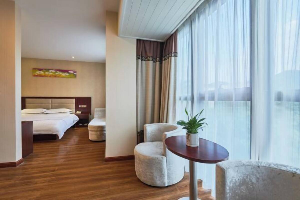 A hotel room with a bed, a round table with a plant, a chair, and a large window with curtains.