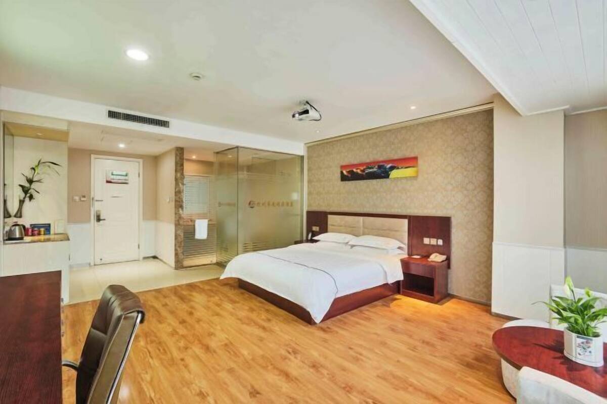 A modern hotel room with a wooden floor, a large bed with white linens, a bedside table with a lamp, and a wall-mounted artwork.