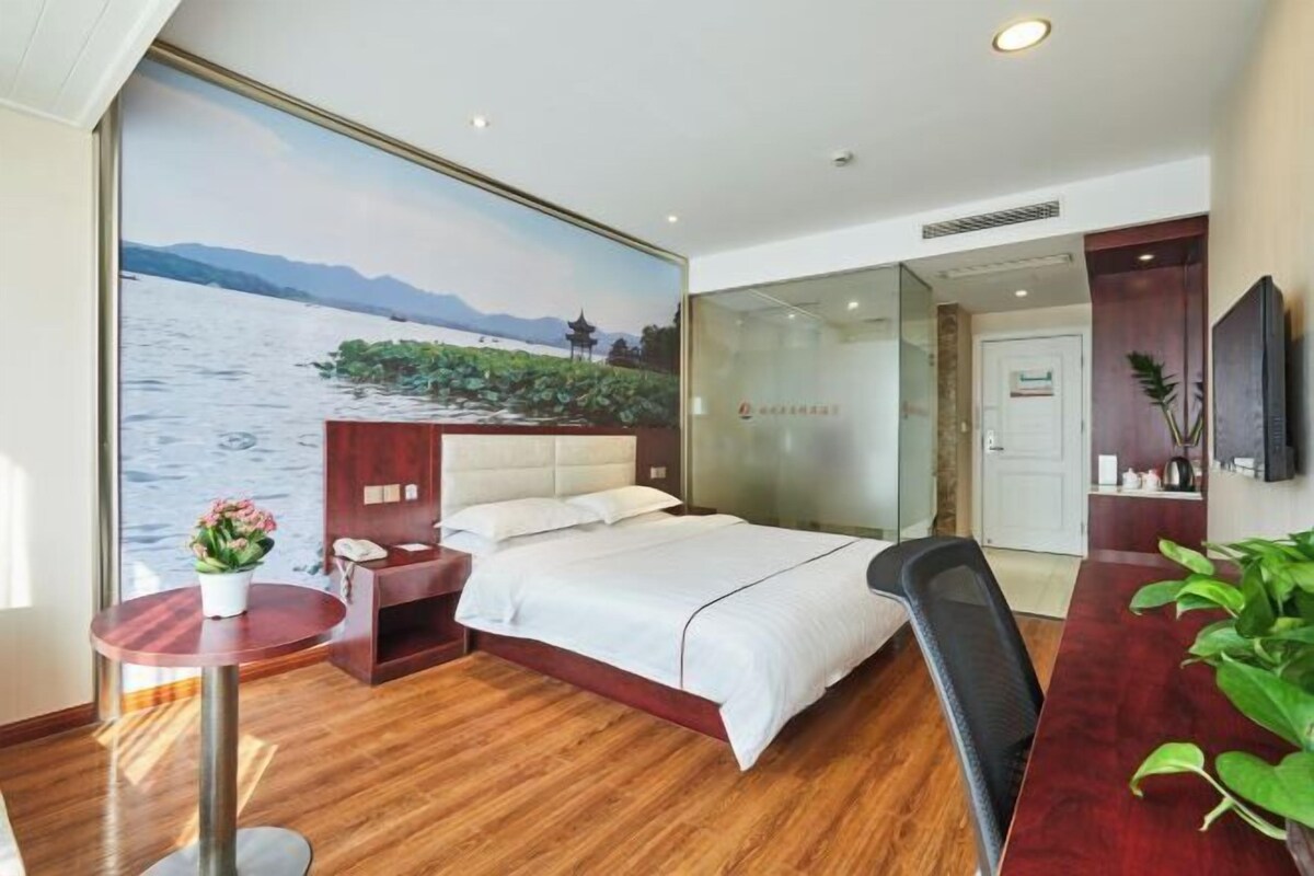 Habitación de hotel con un ventanal grande que ofrece una vista panorámica del agua y las montañas, una cama con sábanas blancas, una mesita redonda con una planta y un escritorio con una silla.