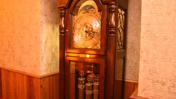 A wooden clock with a detailed face and pendulum, set in a room with wooden paneling and a textured wall.