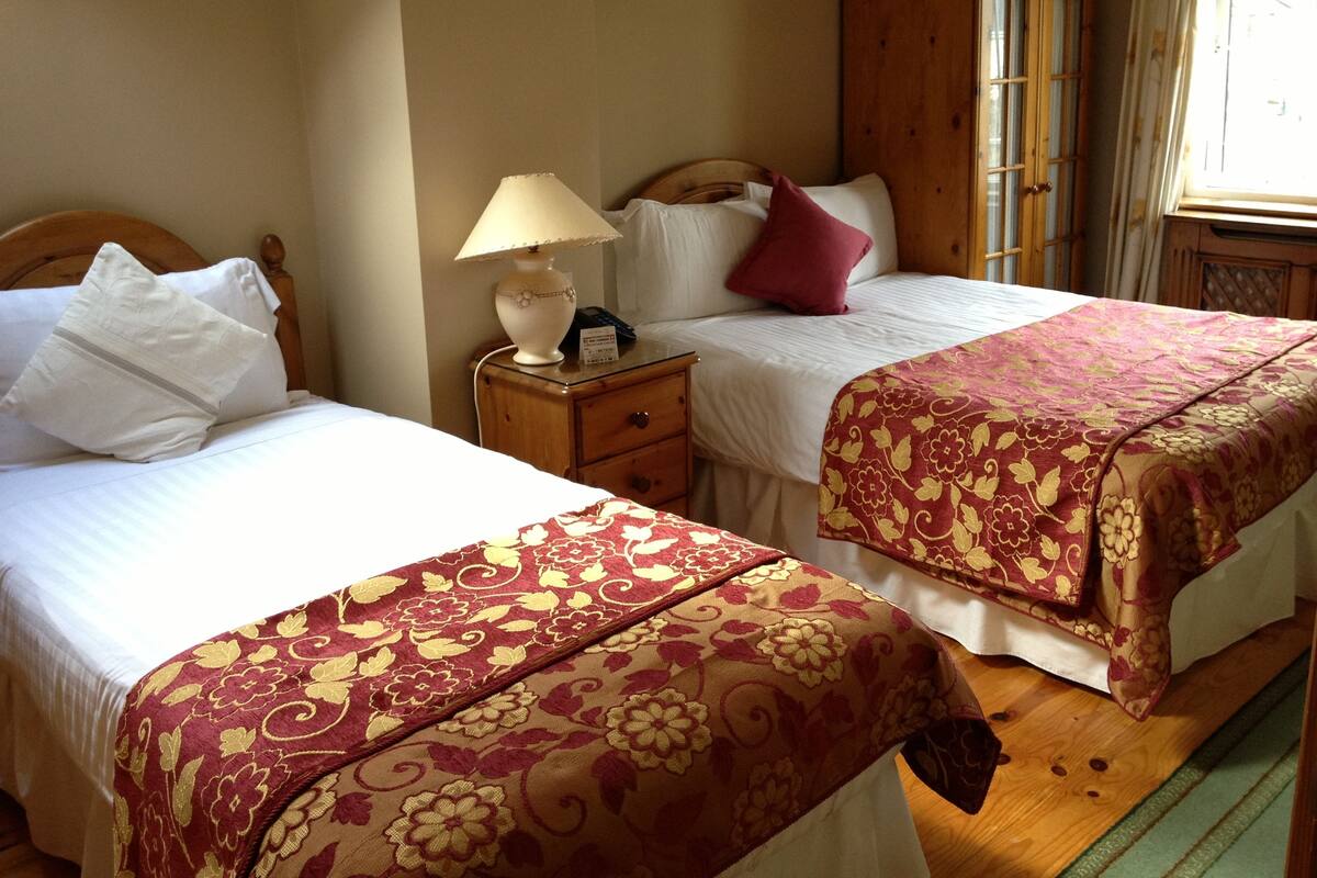 A bedroom with two beds, wooden headboards, a nightstand with a lamp, and a window with curtains.
