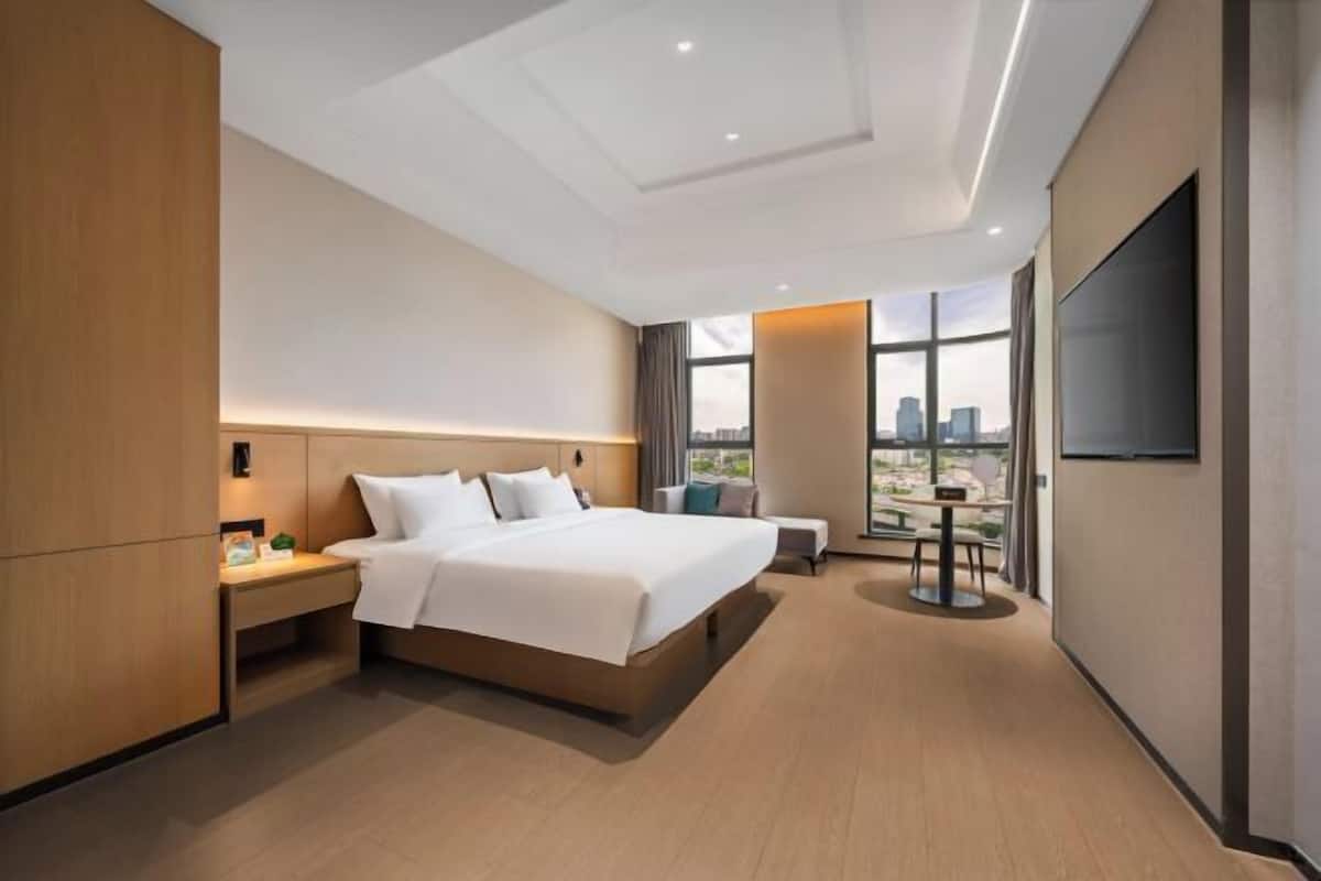 A modern hotel room with a large bed, a desk, a chair, a TV, and a city view.
