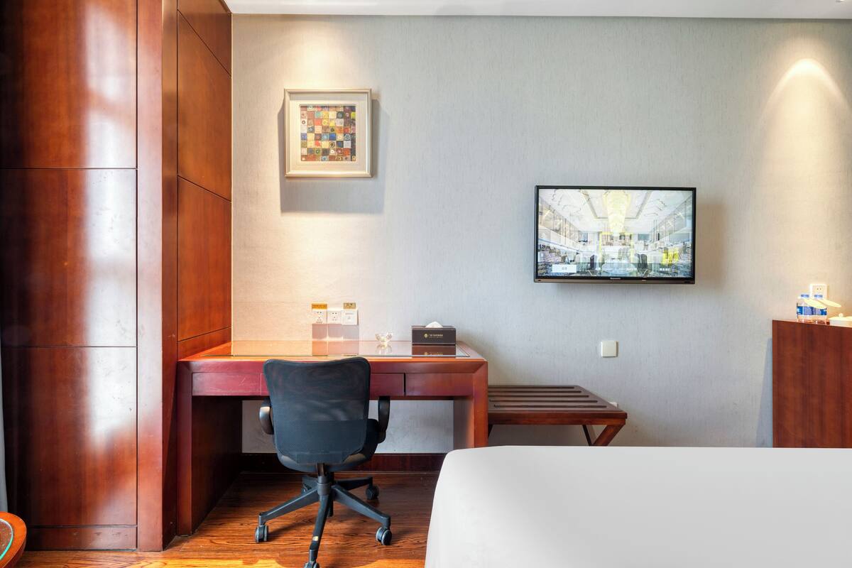 A hotel room with a wooden desk, a black office chair, a bed, a flat-screen TV mounted on the wall, and a framed picture above the desk.