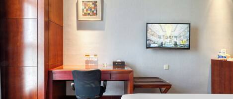 A hotel room with a wooden desk, a black office chair, a bed, a flat-screen TV mounted on the wall, and a framed picture above the desk.
