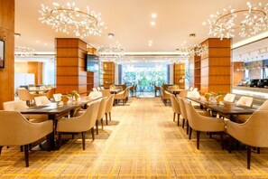 Comedor de hotel con varias mesas y sillas, alfombra estampada y luminarias decorativas.