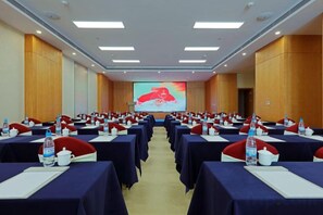 Un salón de conferencias con filas de mesas, cada una con una botella de agua, una taza blanca y un cojín rojo.