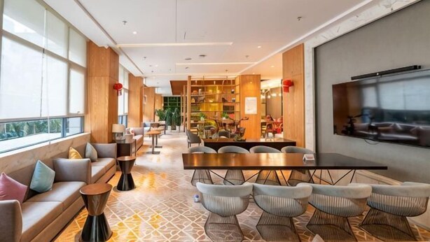 Un hall d’hôtel spacieux avec une longue table à manger, des sièges confortables, un téléviseur fixé au mur et de grandes fenêtres.