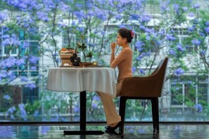 A person seated at a table with a view of purple flowers through a window. - Yunnan Weiju Golden Eagle Grand Hotel (Kunming Railway Station Shuangqiao Night Market Style Street) (Kunming)