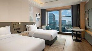 Quarto de hotel com duas camas, uma mesinha, uma televisão, uma mesinha de cabeceira e uma janela grande com vista para a cidade.