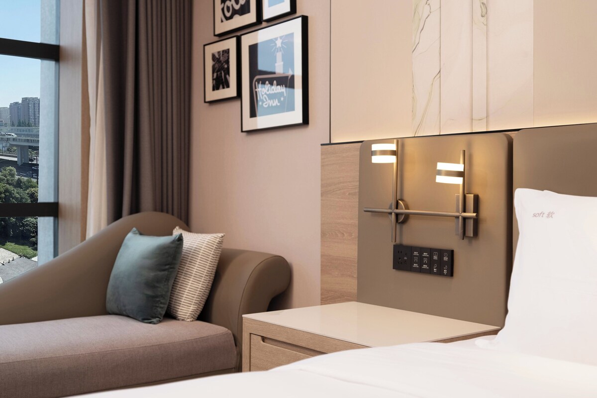 Habitación de hotel con cama, mesita de noche, una silla, ventana con vistas a edificios y cuadros en la pared.