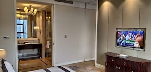 A modern hotel room with a large TV, a bathroom with a mirror and sink, and a wooden cabinet.