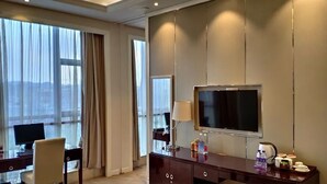 A hotel room with a large window, a desk with a computer, a TV, and a minibar.