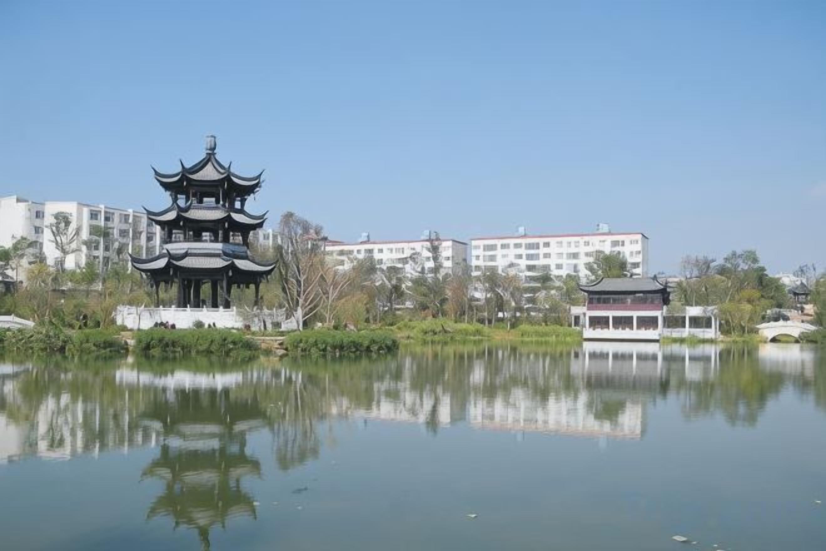 Perinteinen pagodityylinen rakennus, joka heijastuu tyynen vesialueen pinnasta. Sen ympärillä on moderneja rakennuksia ja vehreää kasvillisuutta.