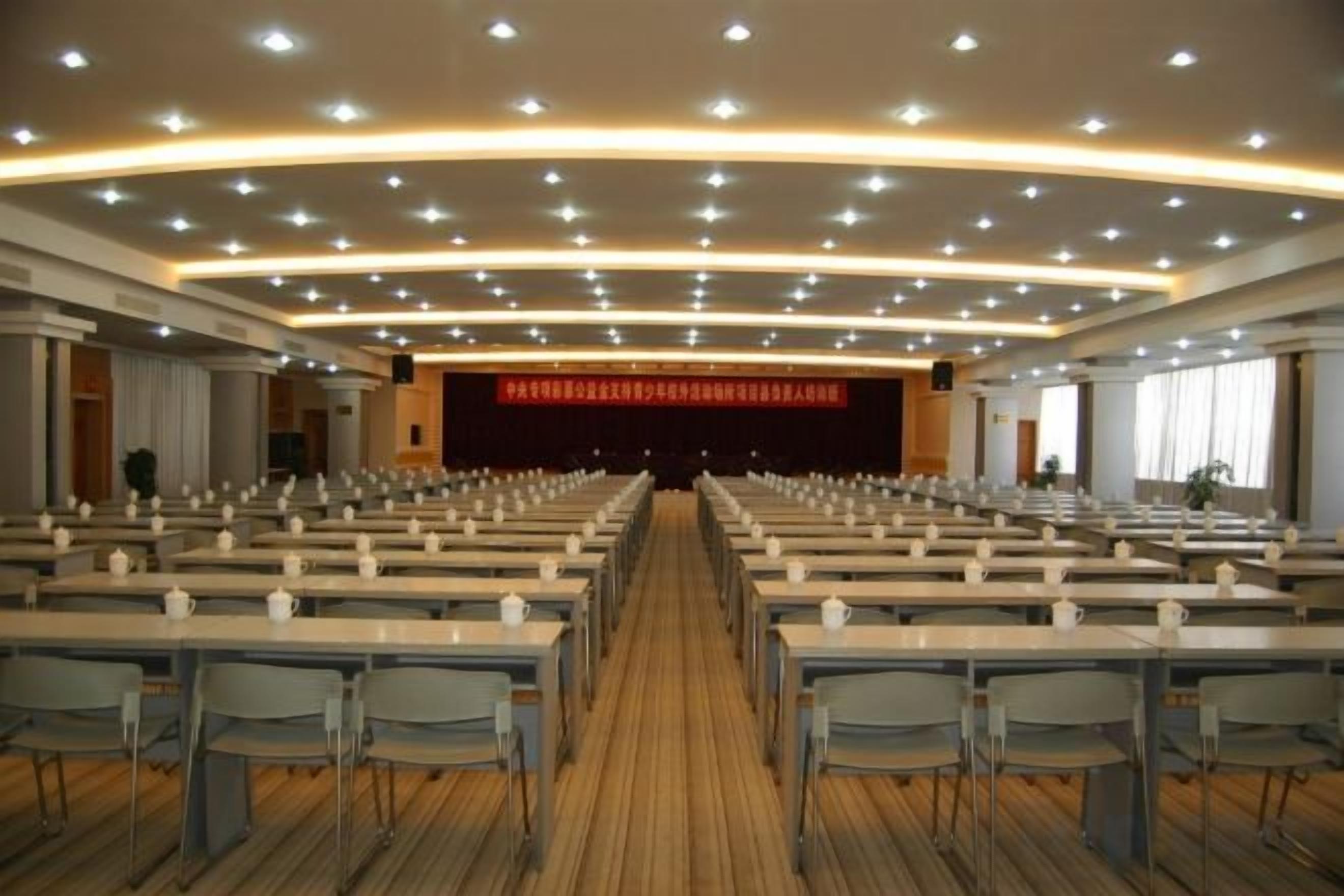 A conference room with rows of tables and chairs, a stage with a red backdrop, and a wooden floor.