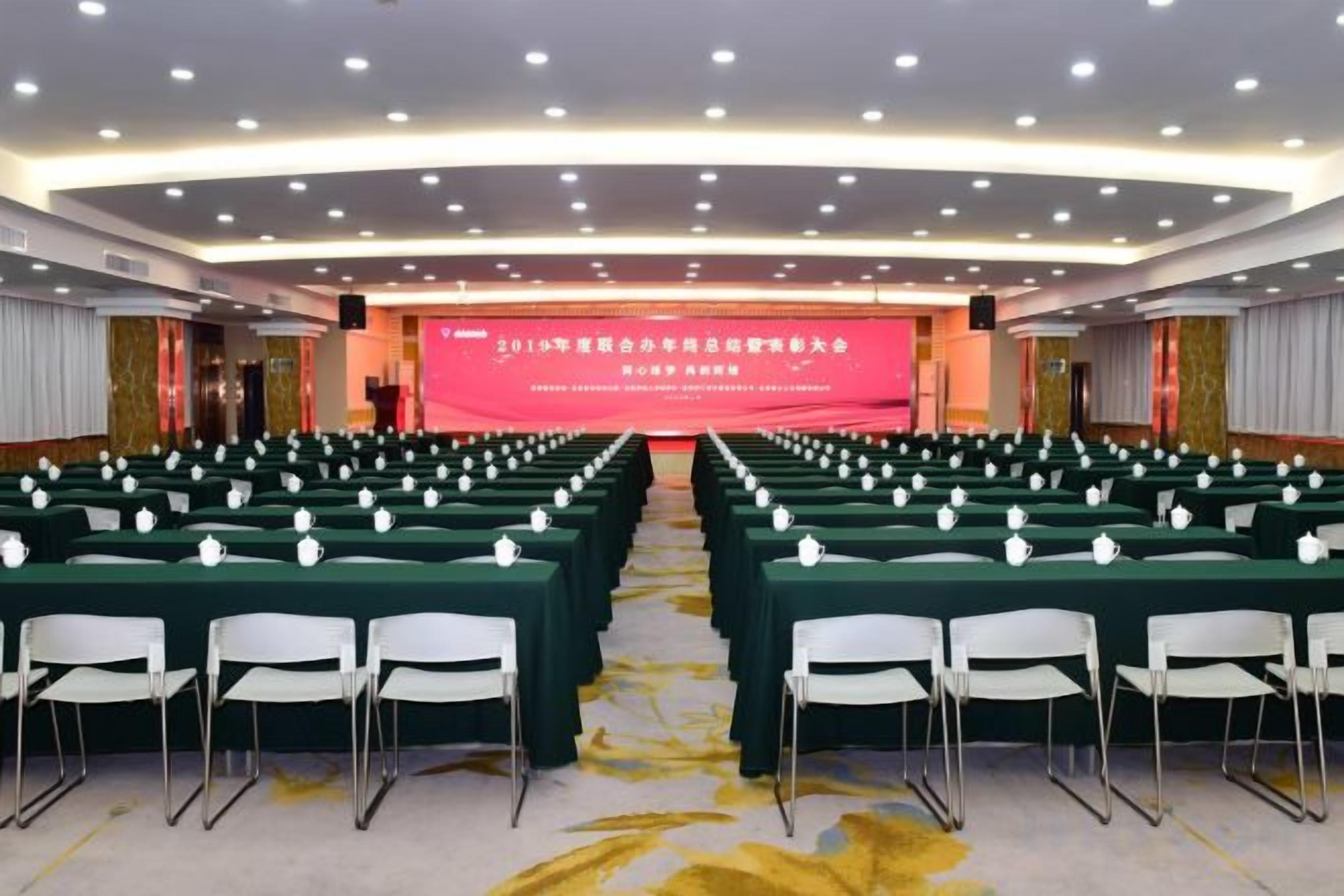 A conference room with rows of tables and chairs, a stage with a red backdrop, and a modern ceiling design.