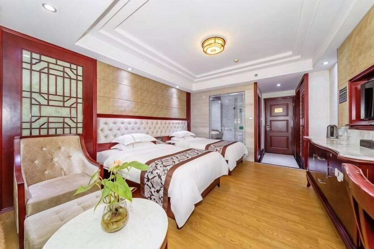 Une chambre d’hôtel avec un grand lit, un canapé, une petite table avec un vase, et une salle de bain visible à travers une porte ouverte.