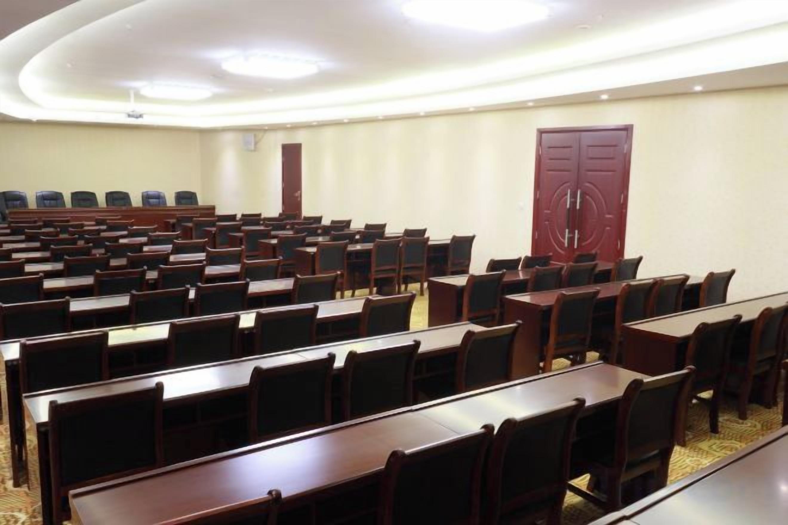 A conference room with rows of tables and chairs, a large red door, and a carpeted floor.