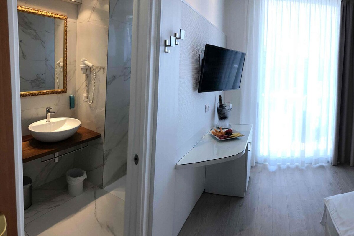 Ein Hotelzimmer mit einem Bett, einem Nachttisch mit Telefon, einem Sessel, einem an der Wand montierten Fernseher, einem Badezimmer mit Waschbecken und Spiegel sowie einem großen Fenster mit Vorhängen.