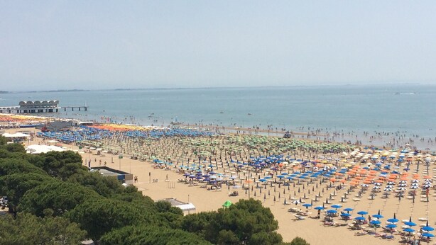 Ein Strand mit vielen Sonnenschirmen und Liegestühlen, ein Steg im Hintergrund und freie Sicht auf das Meer.