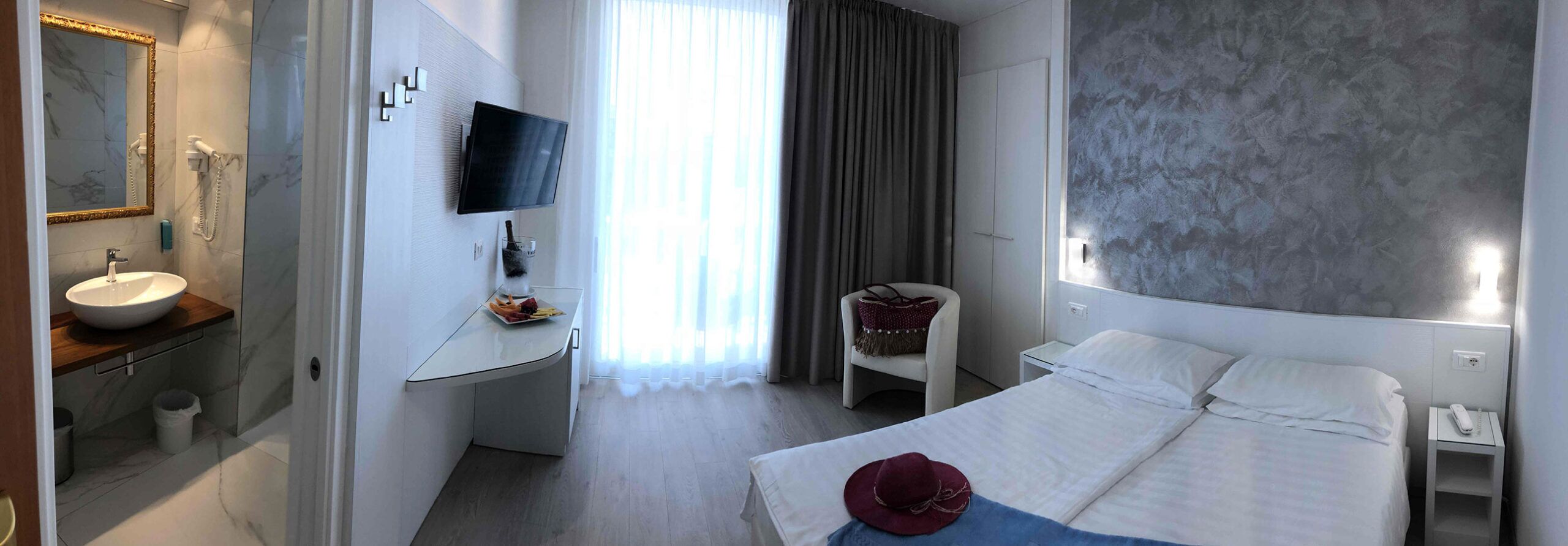A hotel room with a bed, a bedside table with a phone, a chair, a TV mounted on the wall, a bathroom with a sink and mirror, and a large window with curtains.