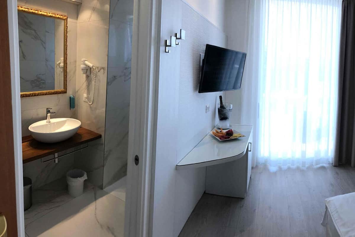 Une chambre d’hôtel comprenant un lit, une table de chevet avec un téléphone, une chaise, un téléviseur fixé au mur, une salle de bain avec un lavabo et un miroir, et une grande fenêtre avec des rideaux.
