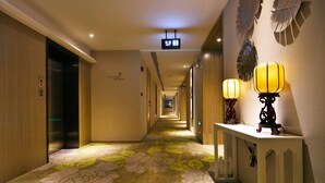 Un pasillo de hotel con alfombra floral, un ascensor, un mostrador de recepción con lámparas y un cartel en la pared.