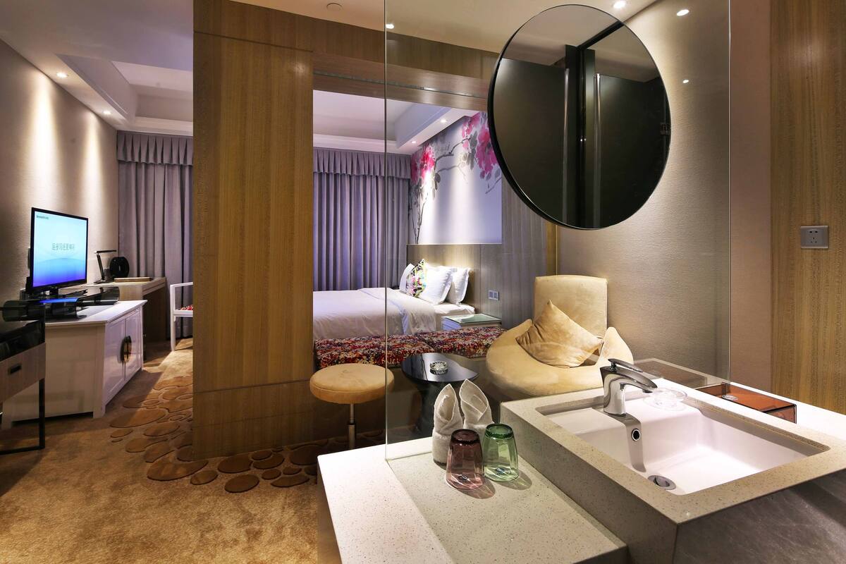 Habitación de hotel moderna con una cama grande, un escritorio con computadora, un baño con lavamanos doble y un espejo grande.