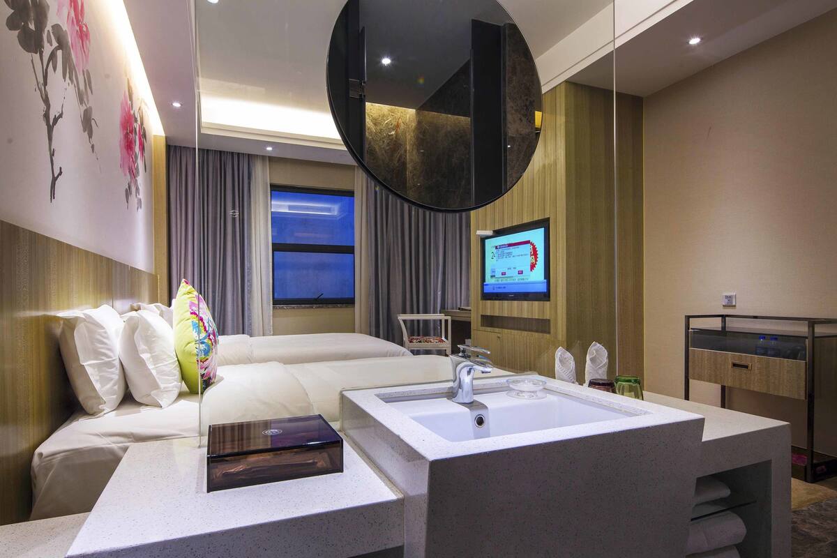 Habitación de hotel con cama, baño con lavamanos, espejo y televisor en la pared.