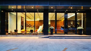 Una entrada de hotel moderna con amplias puertas de vidrio, un mostrador de recepción y una zona de estar con sillas y una mesa.
