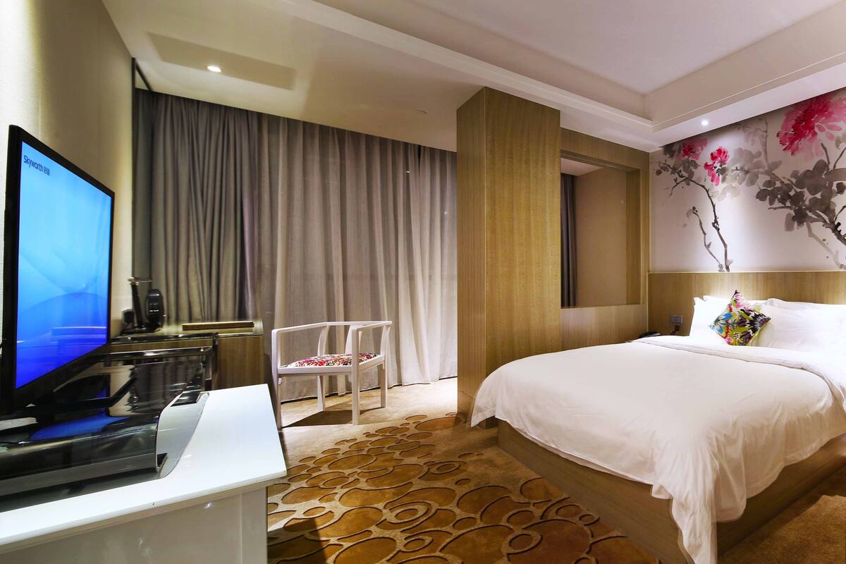 Una habitación de hotel con una cama, un escritorio con una computadora, una silla, un televisor y un cuadro floral en la pared.