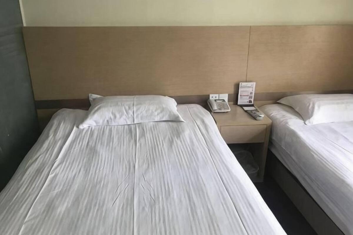 Two single beds with white linens, a wooden headboard, a bedside table with a phone and a sign, and a wall-mounted light.